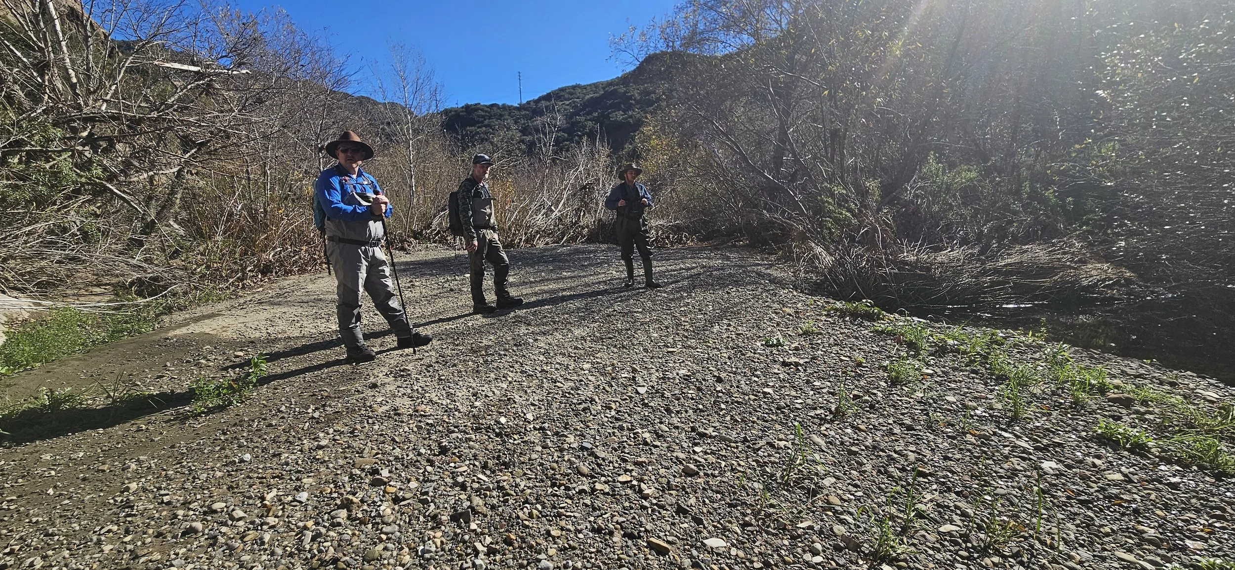 Three people in outdoor gear standing on a gravel path surrounded by trees and mountains.