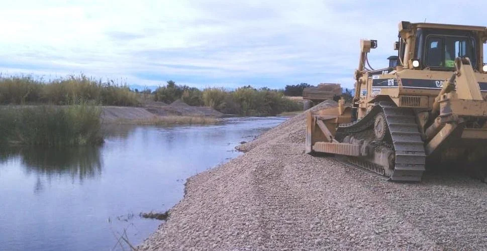 Bulldozer working on riverbank construction with water and vegetation in the background.