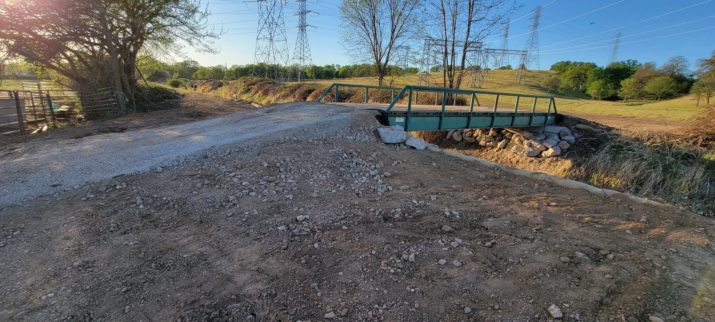 Dirt road leading to a small metal bridge in a rural area, with utility towers and green fields in the background.