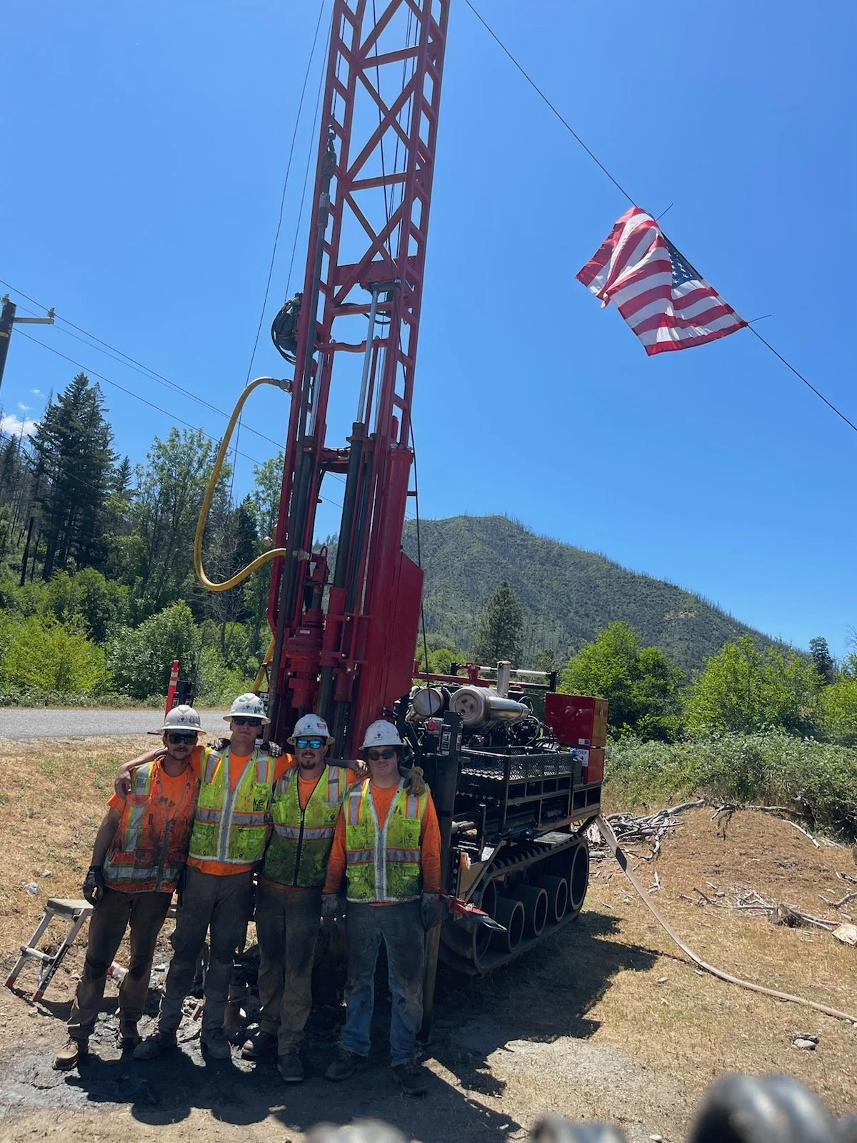 Five construction workers in safety gear standing next to a drilling rig with an American flag overhead, in a rural outdoor setting.