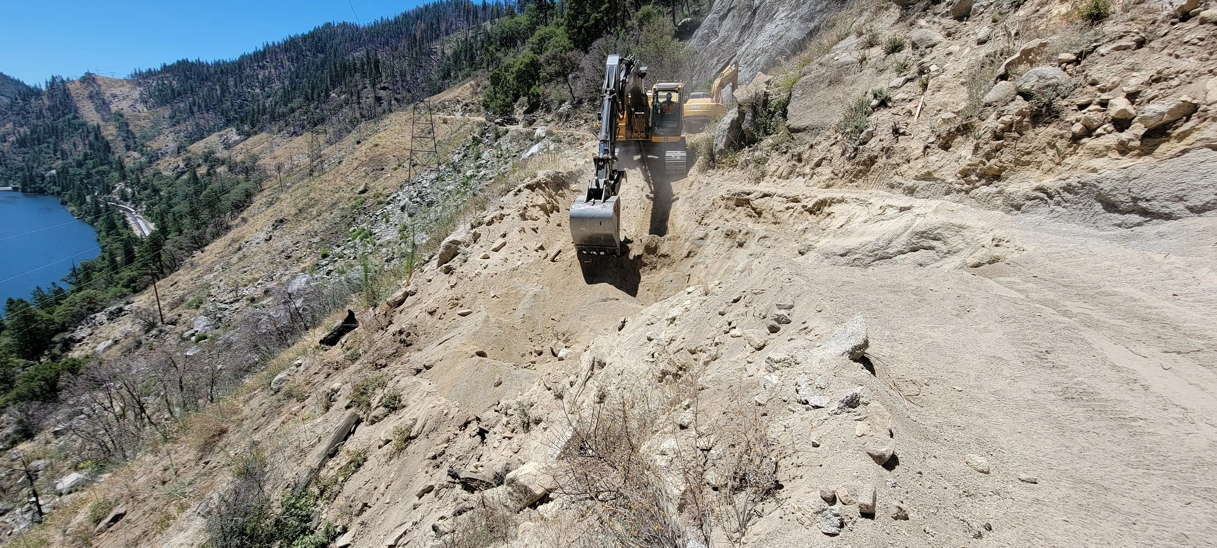 Excavator working on a mountainous terrain with a lake and trees in the background.