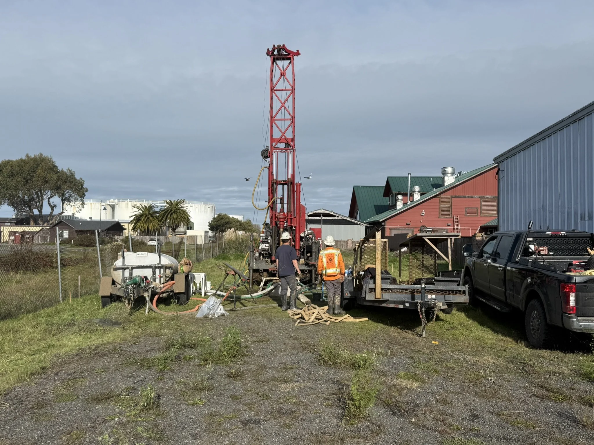 Construction site with drilling equipment and workers, red drilling rig, vehicles, and buildings in the background.