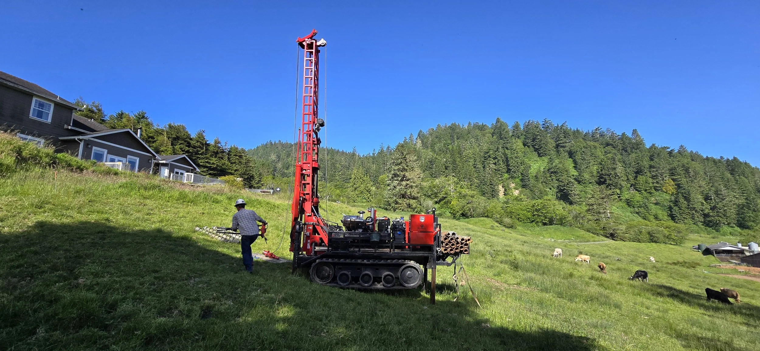 Red drilling rig on grassy hilltop near houses and grazing cattle under clear blue sky.