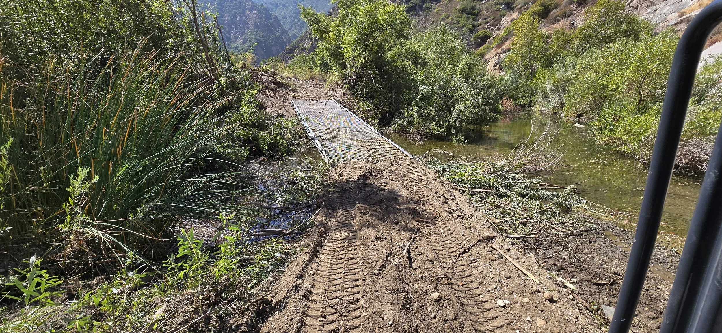 Dirt path with tire tracks leading to a small bridge over a stream, surrounded by green vegetation and hills.