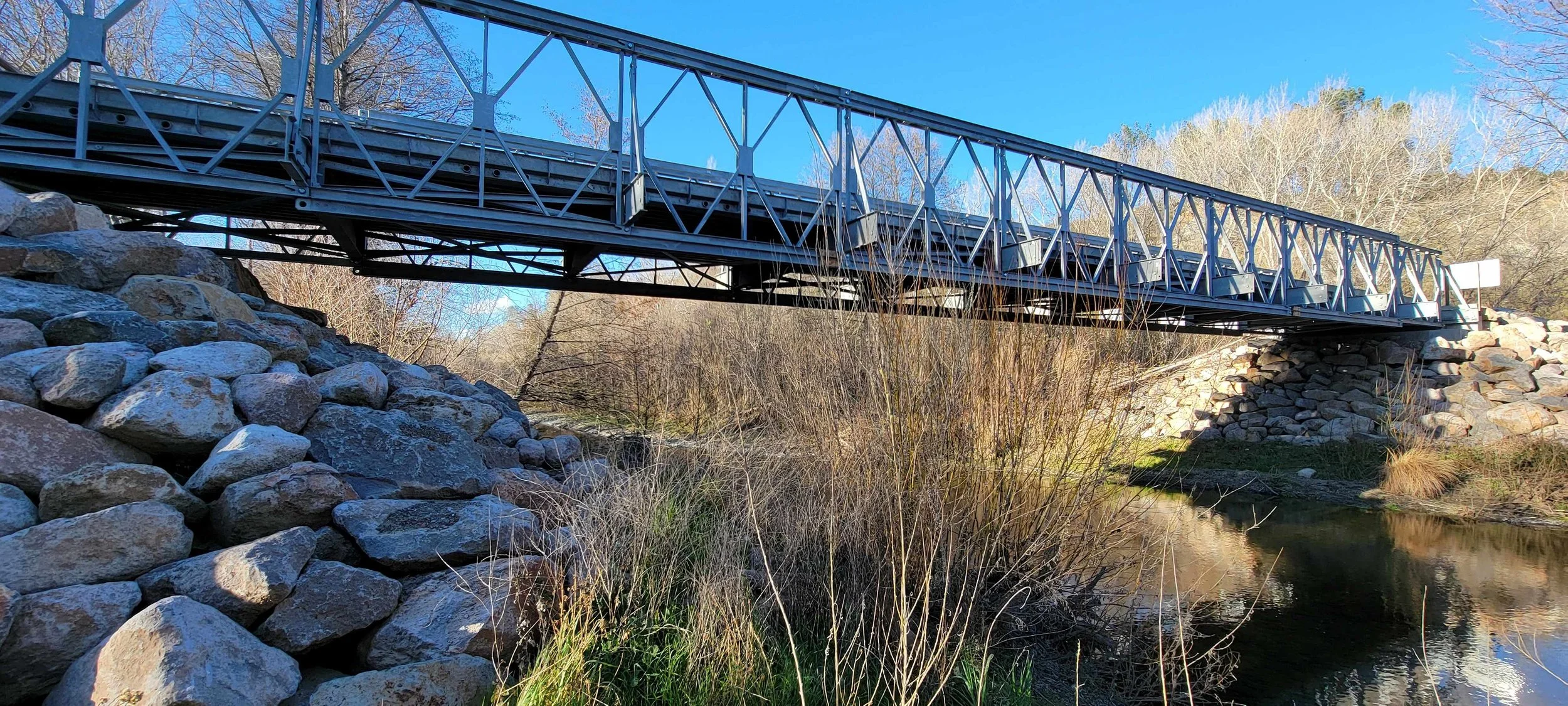 Steel pedestrian bridge over a rocky stream with surrounding trees