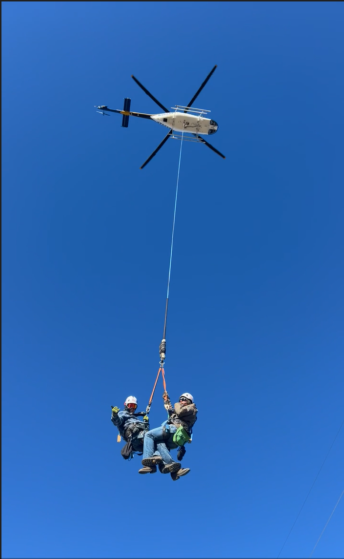Helicopter rescue operation with two people suspended below, clear blue sky.