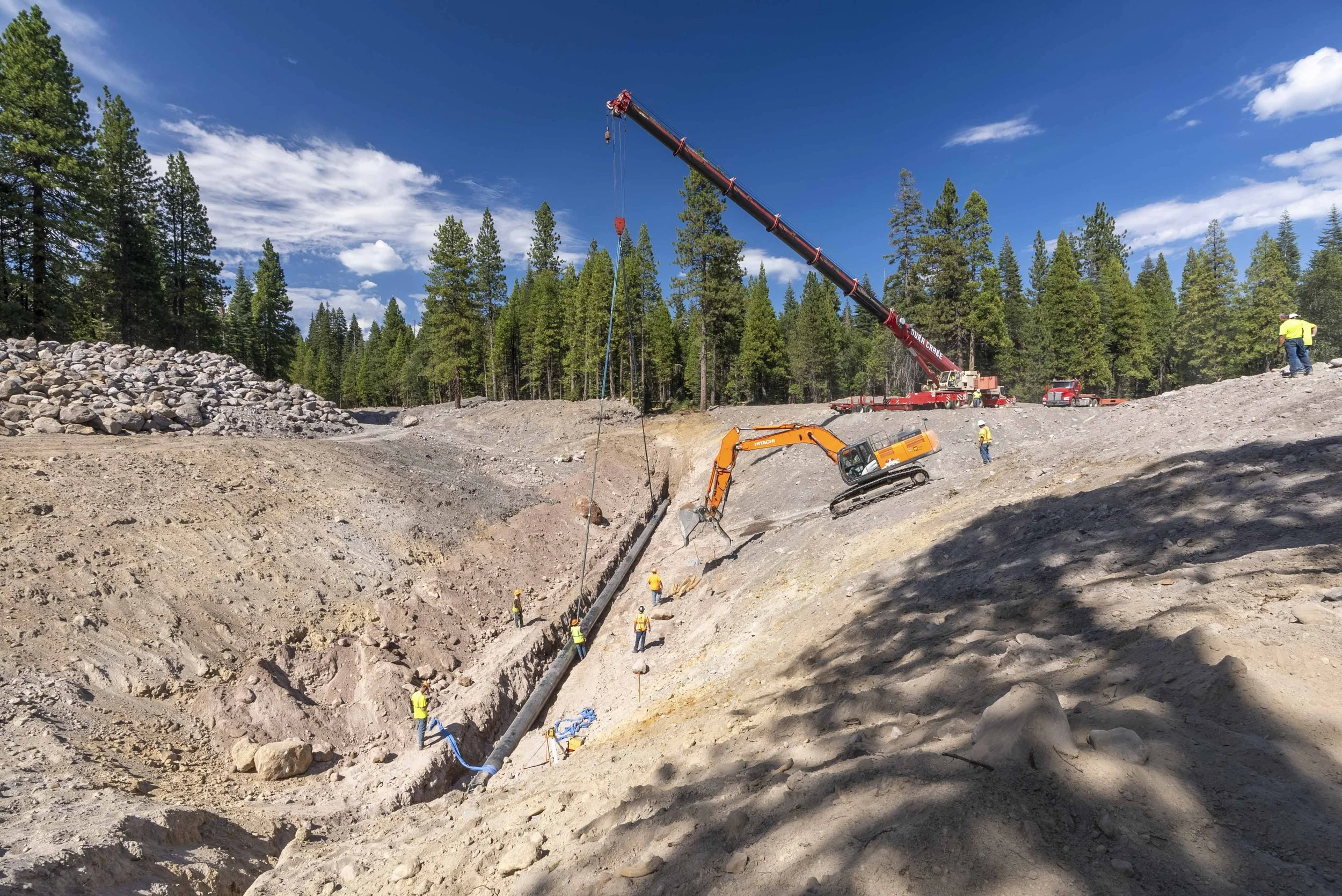 Construction site with heavy machinery and workers in a forested area, showing an excavator, a crane, and people working on a trench.