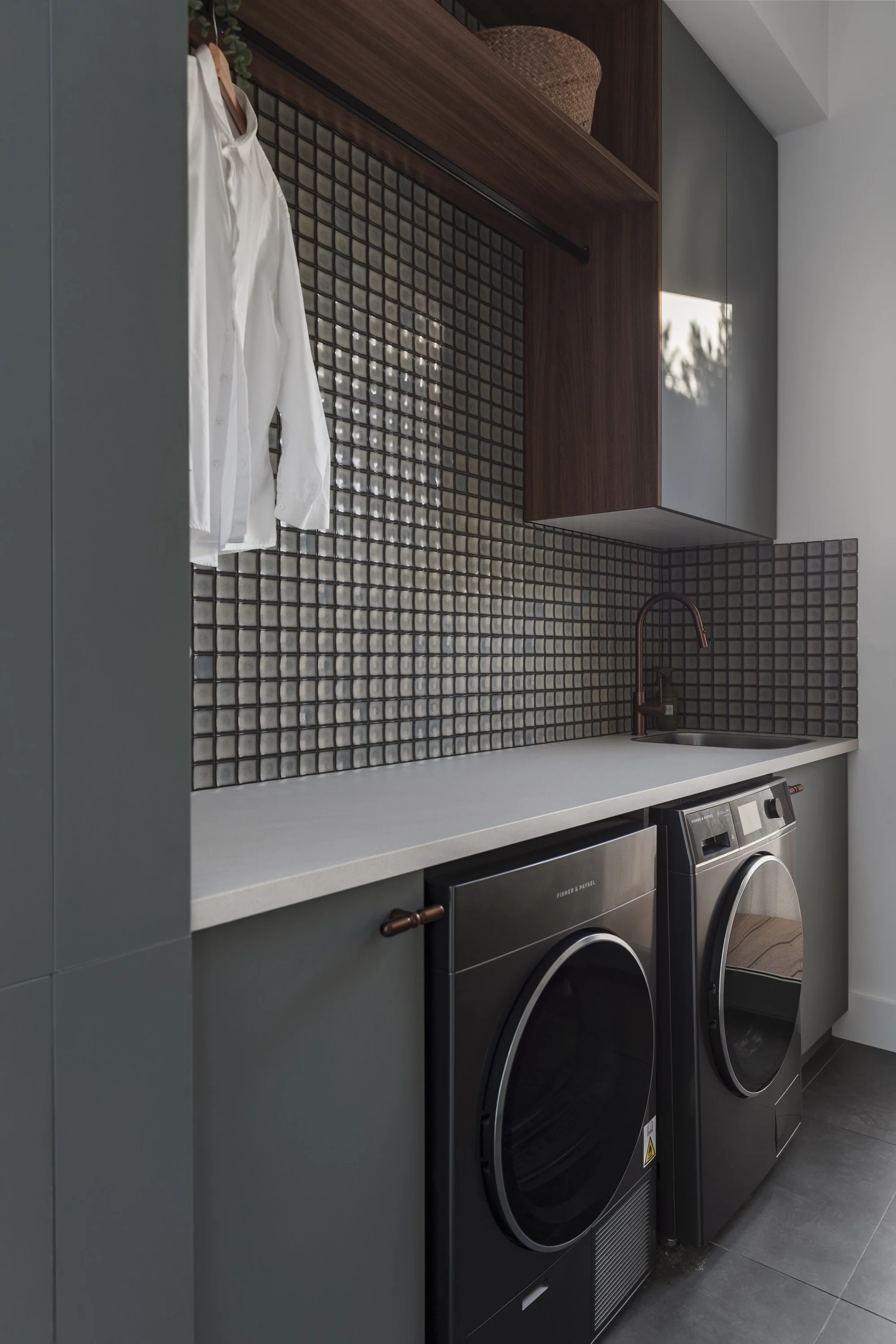 A modern laundry room with a dark gray front-loading washer and dryer set, a white countertop above, a wooden cabinet with a basket and a black hanging rod, and a backsplash with small square black and white tiles.