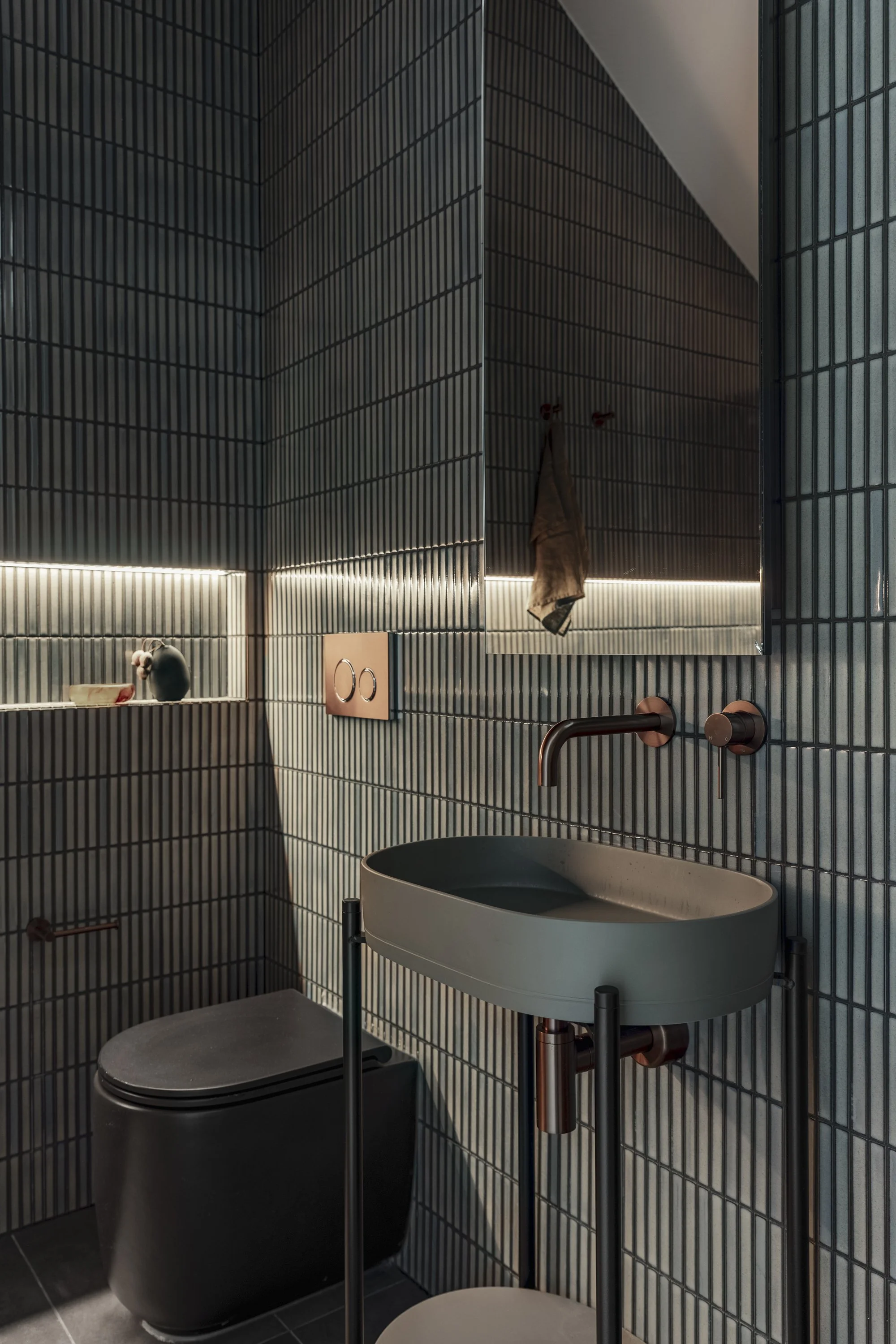 Modern bathroom with black subway tile walls, a wall-mounted mirror, a white oval sink, an industrial-style toilet with a black tank, and copper fixtures, with soft lighting along the wall.