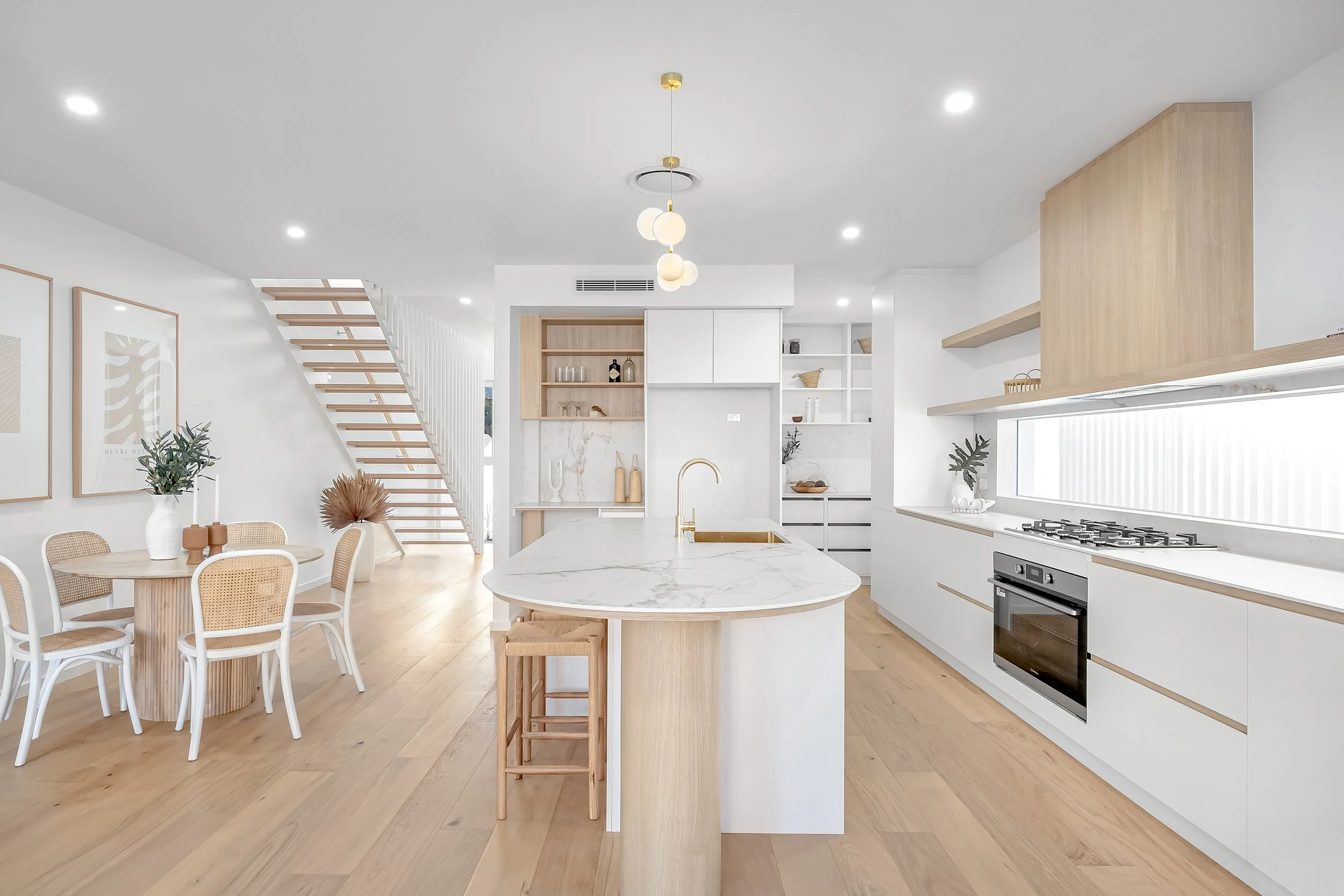 Modern kitchen with white cabinets, marble countertops, a kitchen island with a brass faucet, open wooden shelving, and a window with blinds, adjacent to a dining area with a round table and rattan chairs.