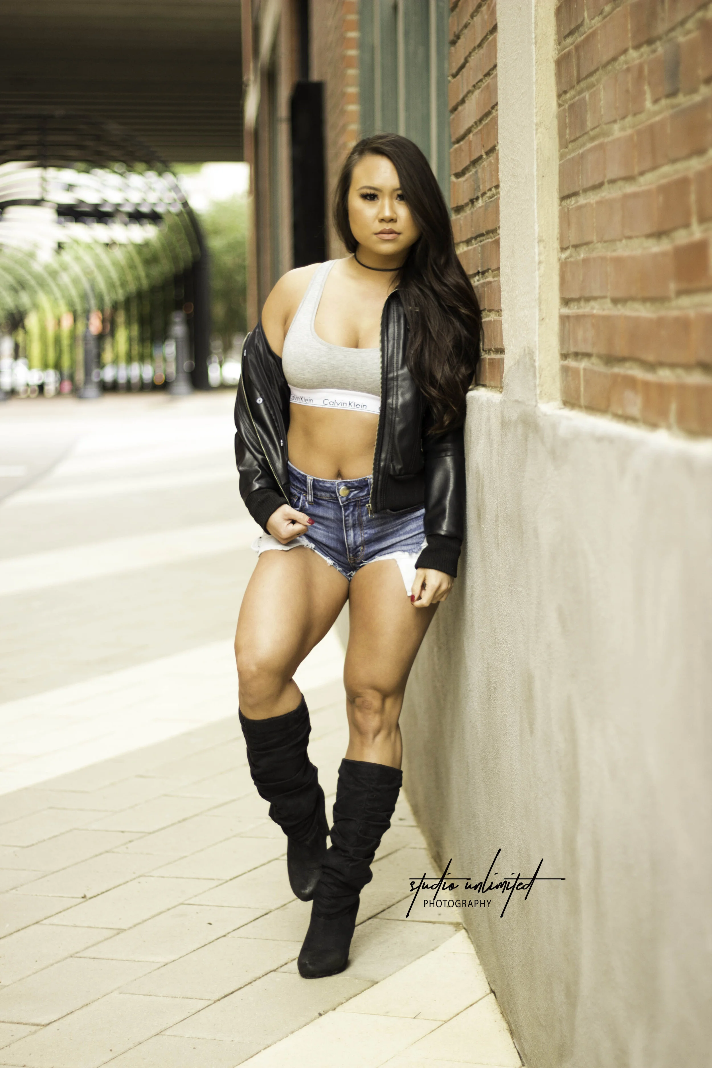 A woman with long dark hair leaning against a brick wall in an urban setting during daytime. She is wearing a gray sports bra, a black leather jacket hanging off her shoulders, denim shorts, and black thigh-high boots. The background shows a sidewalk, green trees, and a covered walkway.