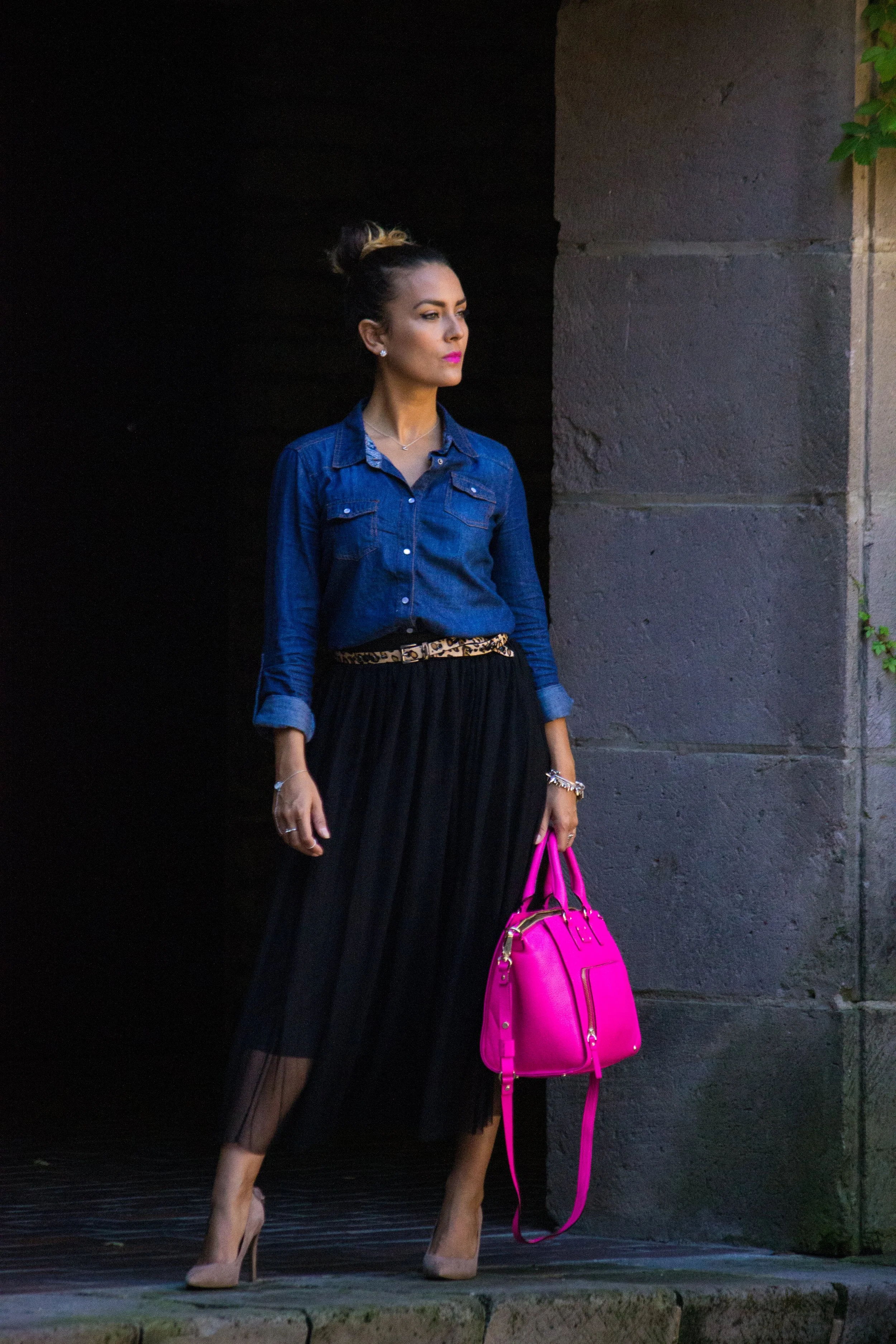A woman standing outdoors against a dark and stone wall background, wearing a denim shirt, black pleated skirt, nude high heels, and carrying a bright pink handbag. Glamour photoshoot