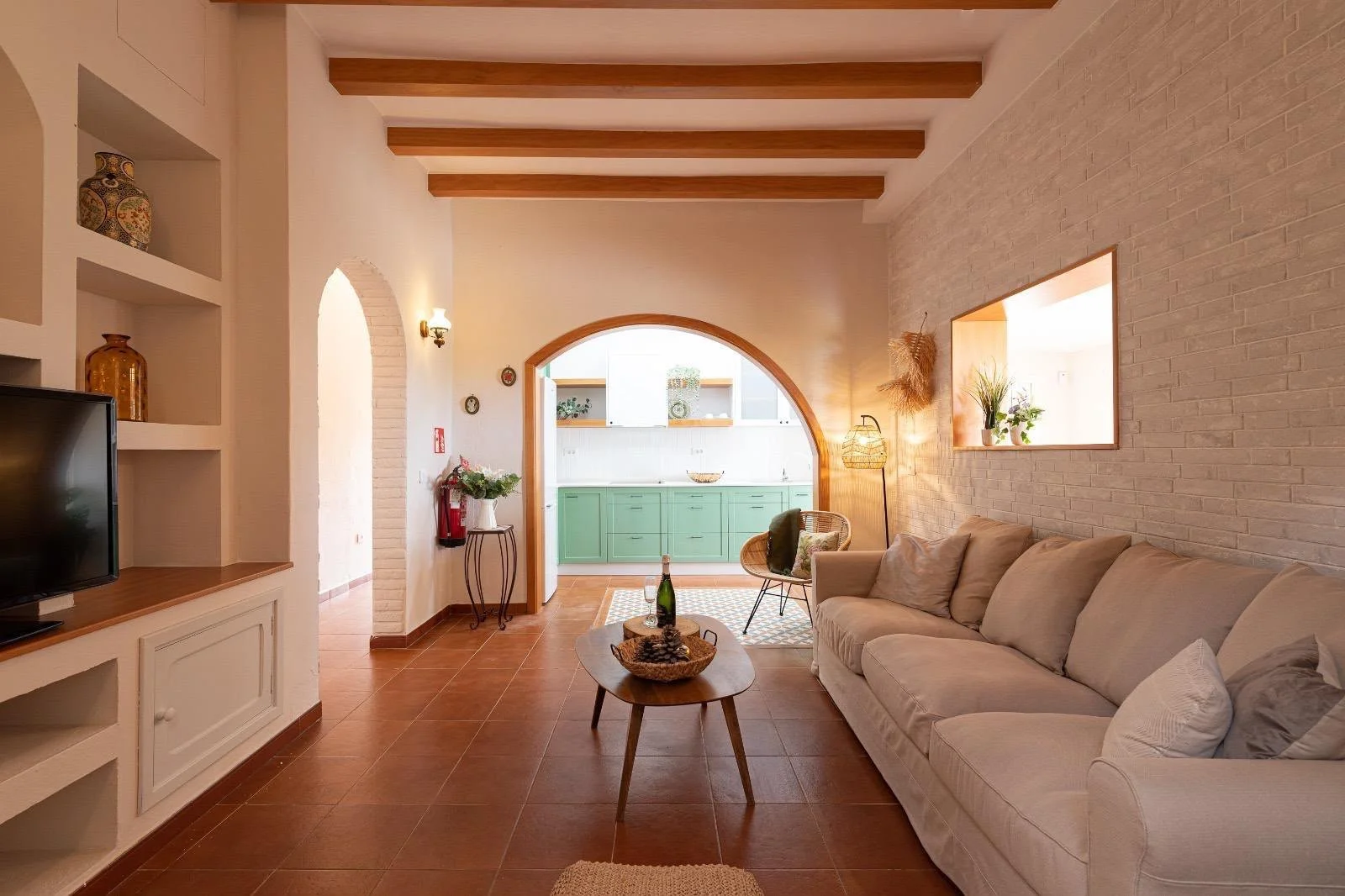 Living room with beige sofa, wooden coffee table with a wine bottle and a bowl, wicker chair, tall floor lamp, decorative wall niche, white brick wall, open kitchen with green cabinets in background, and terracotta floor tiles.