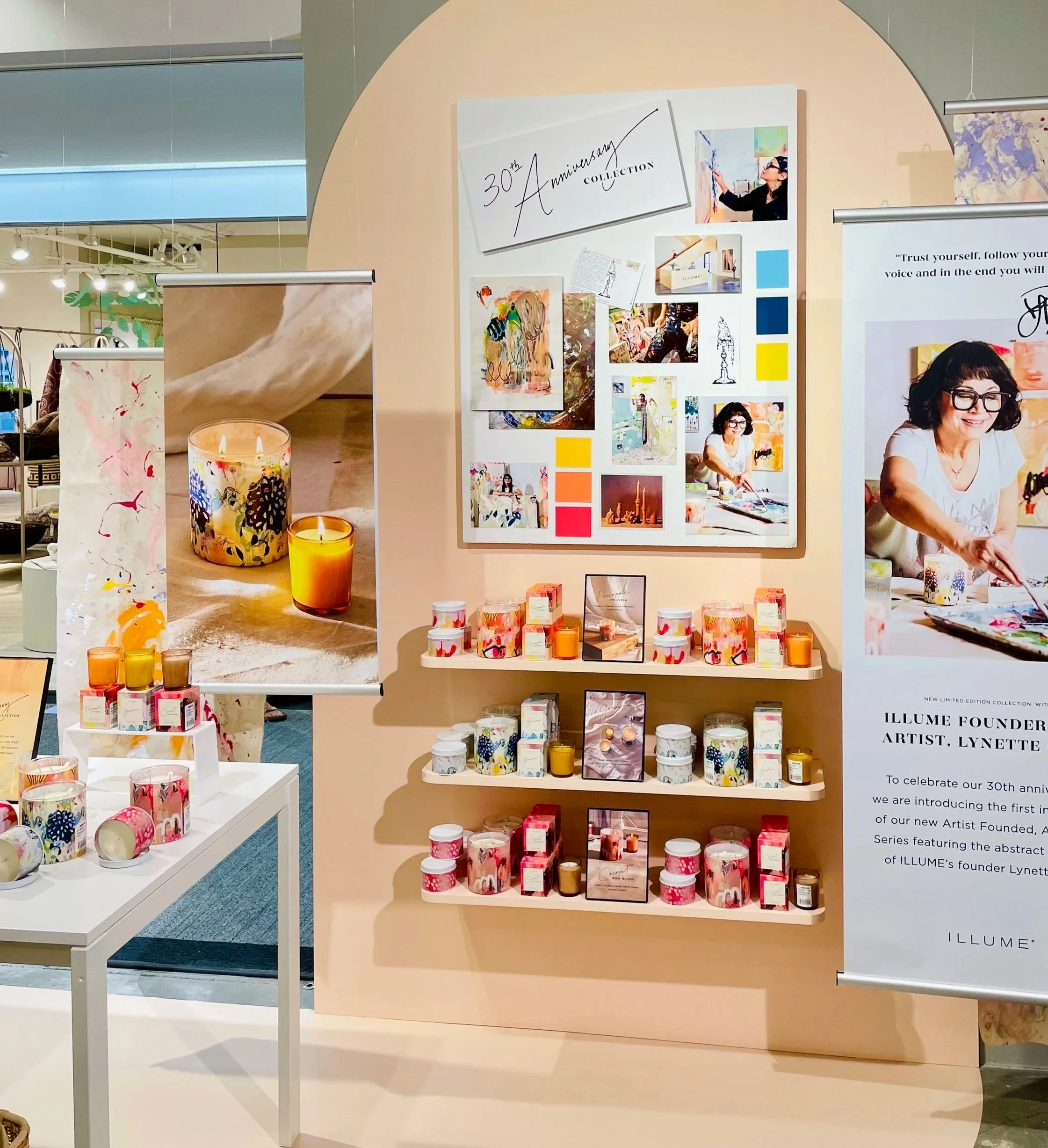 Display of candles, artwork, and information celebrating the 30th anniversary of Illume, a candle company, featuring colorful packaging and a woman painting, in a retail store.
