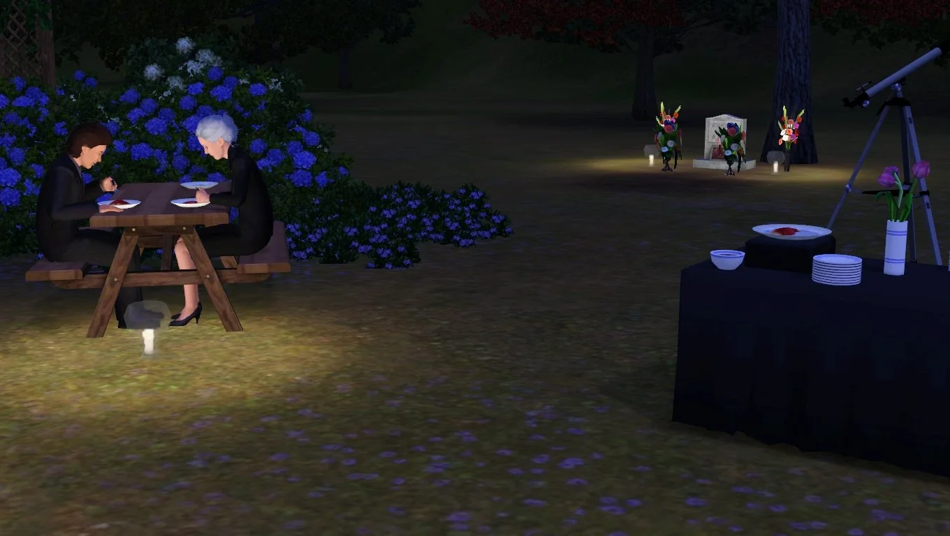 Ryker and Bridget eat at a picnic table at night by candlelight in front of hydrangea bushes. Candles also illuminate a gravestone surrounded by bouquets in the background.