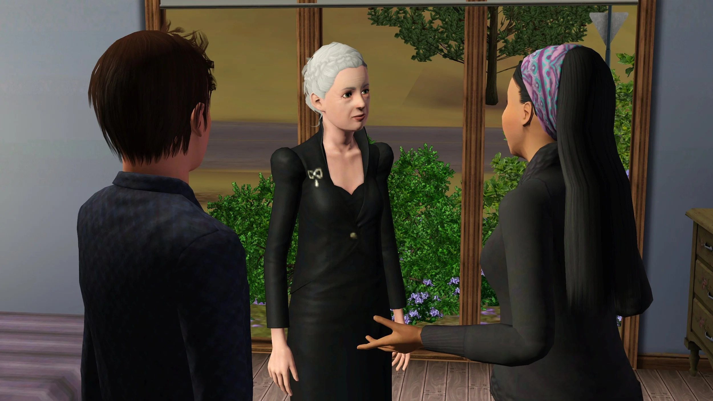 Matthew and Emery stand on either side, facing Bridget. Bridget looks blankly at Emery, who is talking and gesturing with her hands. Bushes, heather and the road are visible through floor-to-ceiling windows behind.