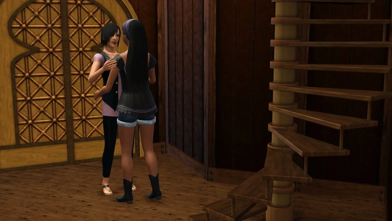 Colette smiles at Emery with a hand on her shoulder & Em's hands near Colette's hips; they stand between Em's front door & spiral staircase. Colette wears a long lilac blouse & long black vest over black leggings; Emery a black blouse & jean shorts.