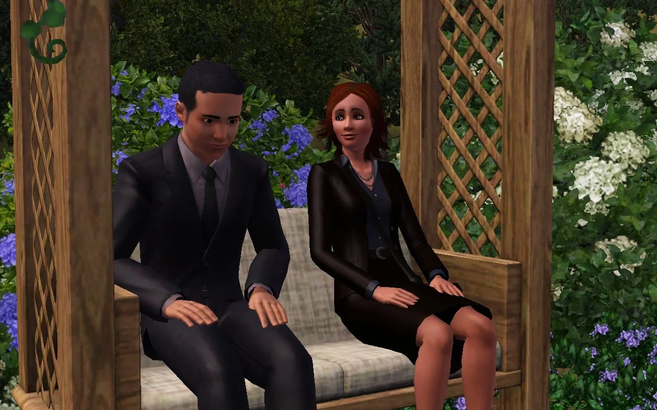 Mason looks towards the ground as he sits next to Alani, who is looking at him with a small smile, on a trellis-covered bench in front of hydrangea bushes. Both have their hands relaxed on their laps and are dressed in dark formalwear.