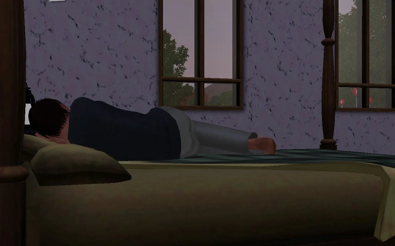 Viewed from behind, Matthew lays in bed on his side, partly curled up, possibly napping. Half the bed is unmade, the bedroom is dark except for early morning light through the windows.