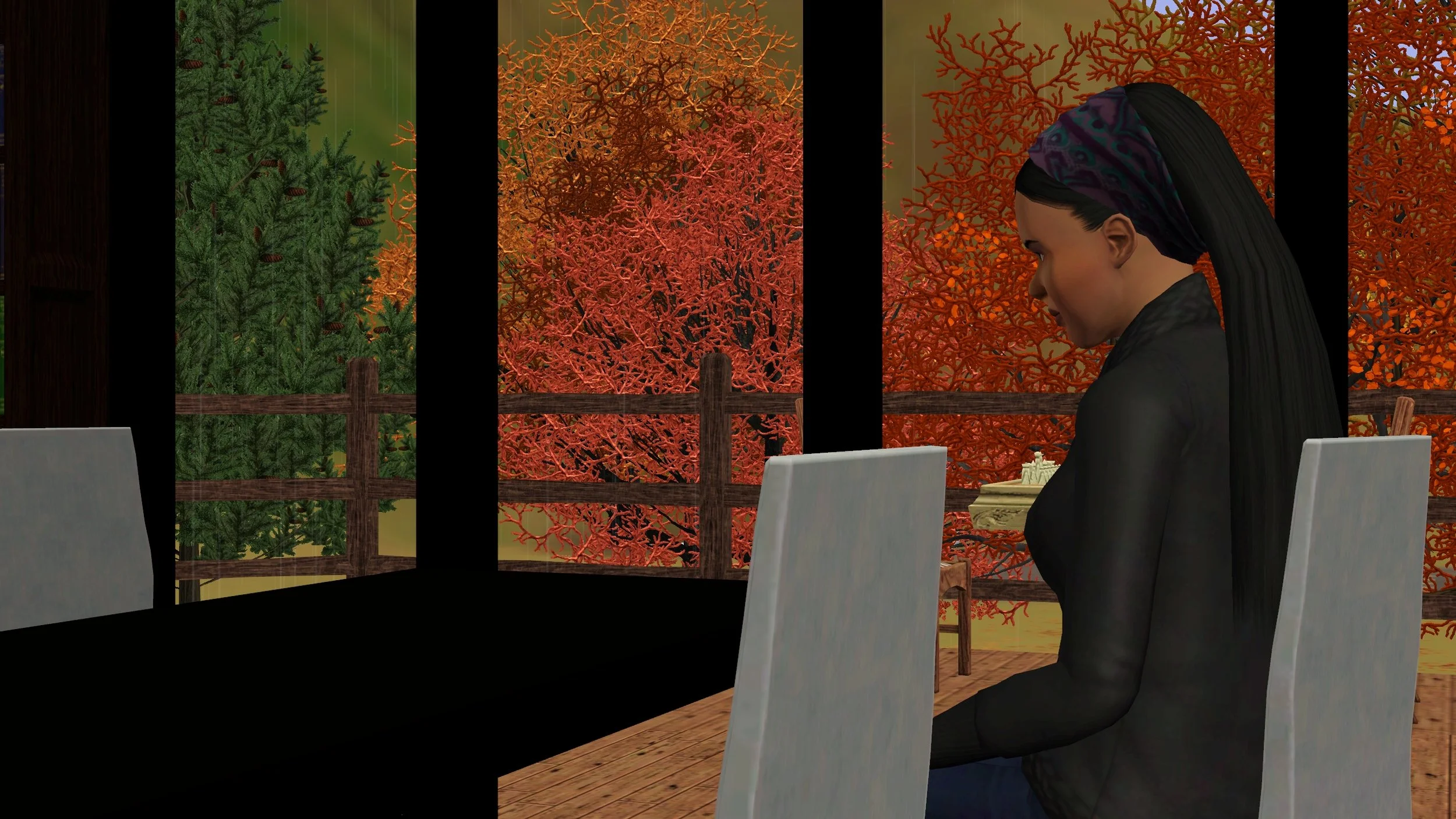 Emery sits in a white wood slab dining chair absently staring at the black dining table in front of her. Four large windows next to her show a wooden deck and numerous colourful autumn trees outside.