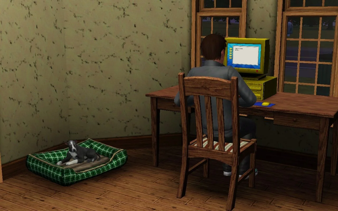 A male Sim sitting at a desk, writing on a computer. A small grey and white dog is lounging in a green plaid dog bed next to the desk.