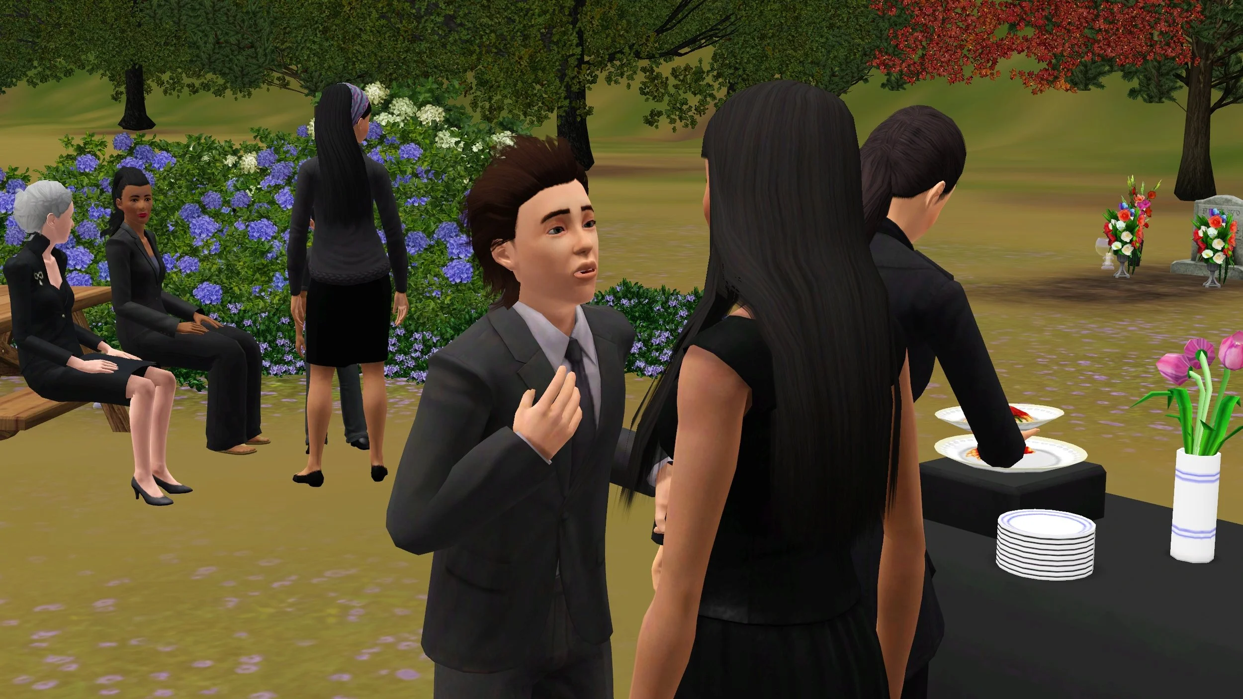 Ryker talks to Sage with a hand near his chest. Behind them: Dorothy takes a serving of spaghetti from a buffet; 4 Sims interact near a picnic table; a gravestone is surrounded by vases of flowers.