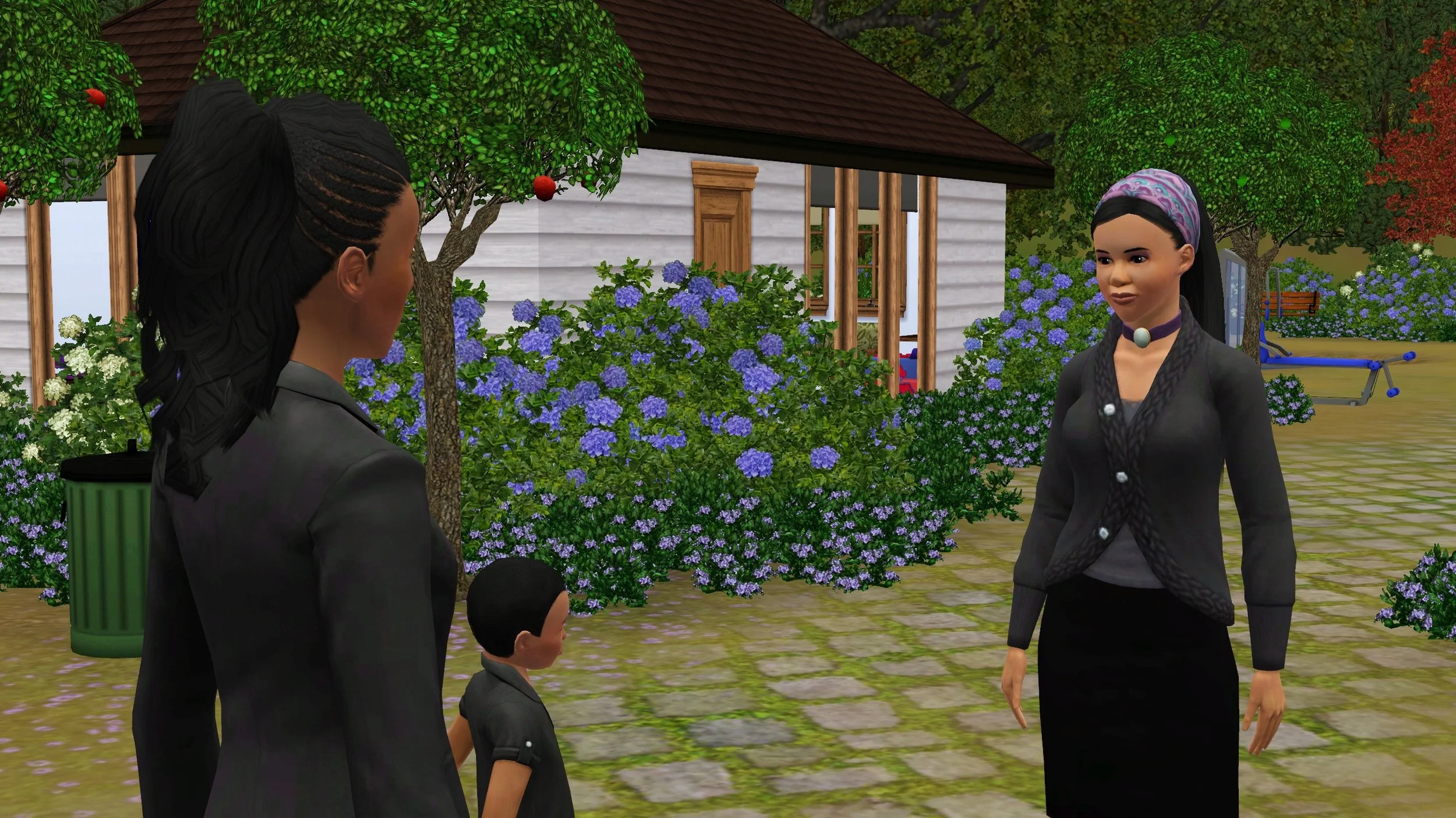 Emery looks down towards Kobe, a small Child Sim standing in front of his mother, Rukiya. Part of the Tevellyn family home is visible behind them, surrounded in blue hydrangeas and purple heather.
