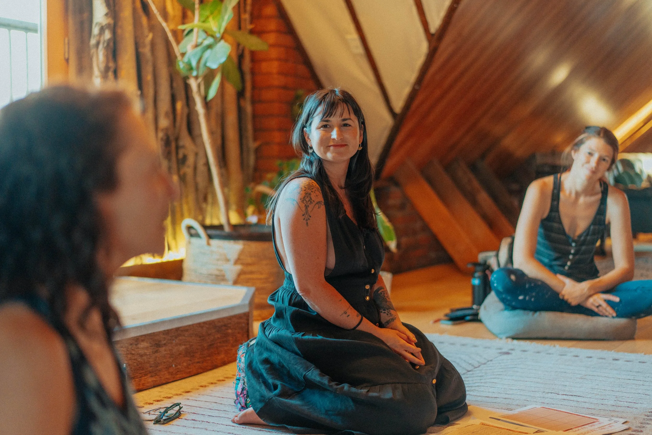 Three women sitting on the floor in a cozy wooden room, with the woman in the middle smiling at the camera.