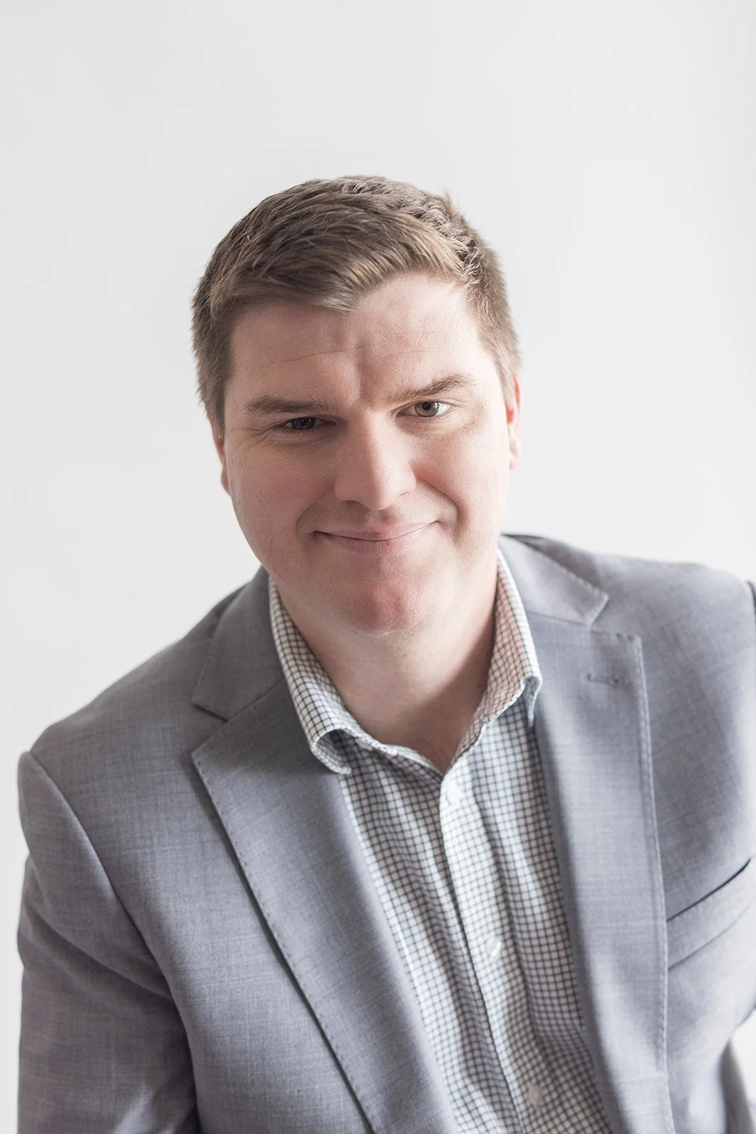 A professional man in a gray suit and checkered shirt, smiling slightly, against a plain white background.