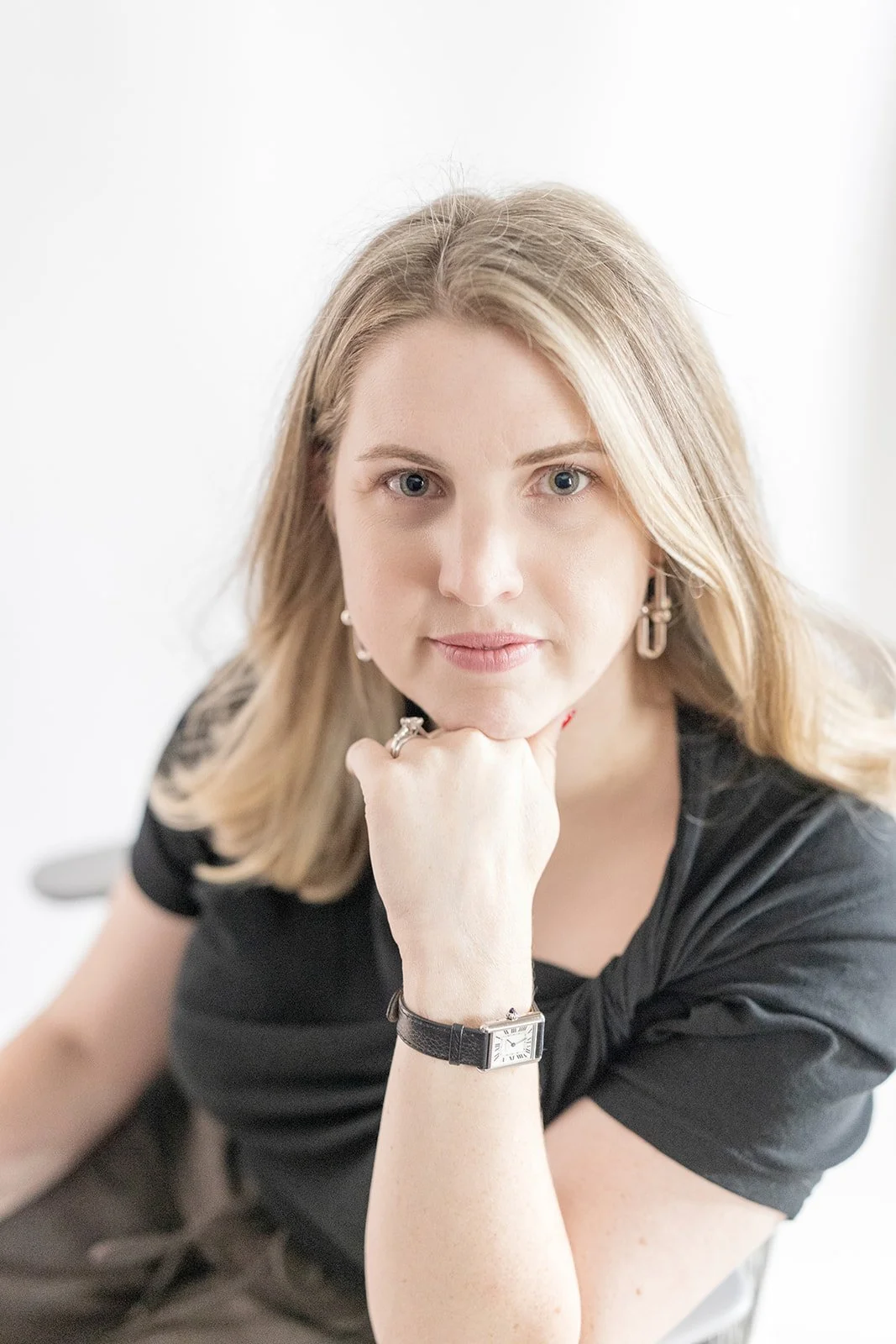 A woman with blonde hair and light skin resting her chin on her hand, wearing a black top, watch, bracelet, earrings, and rings, looking at the camera.