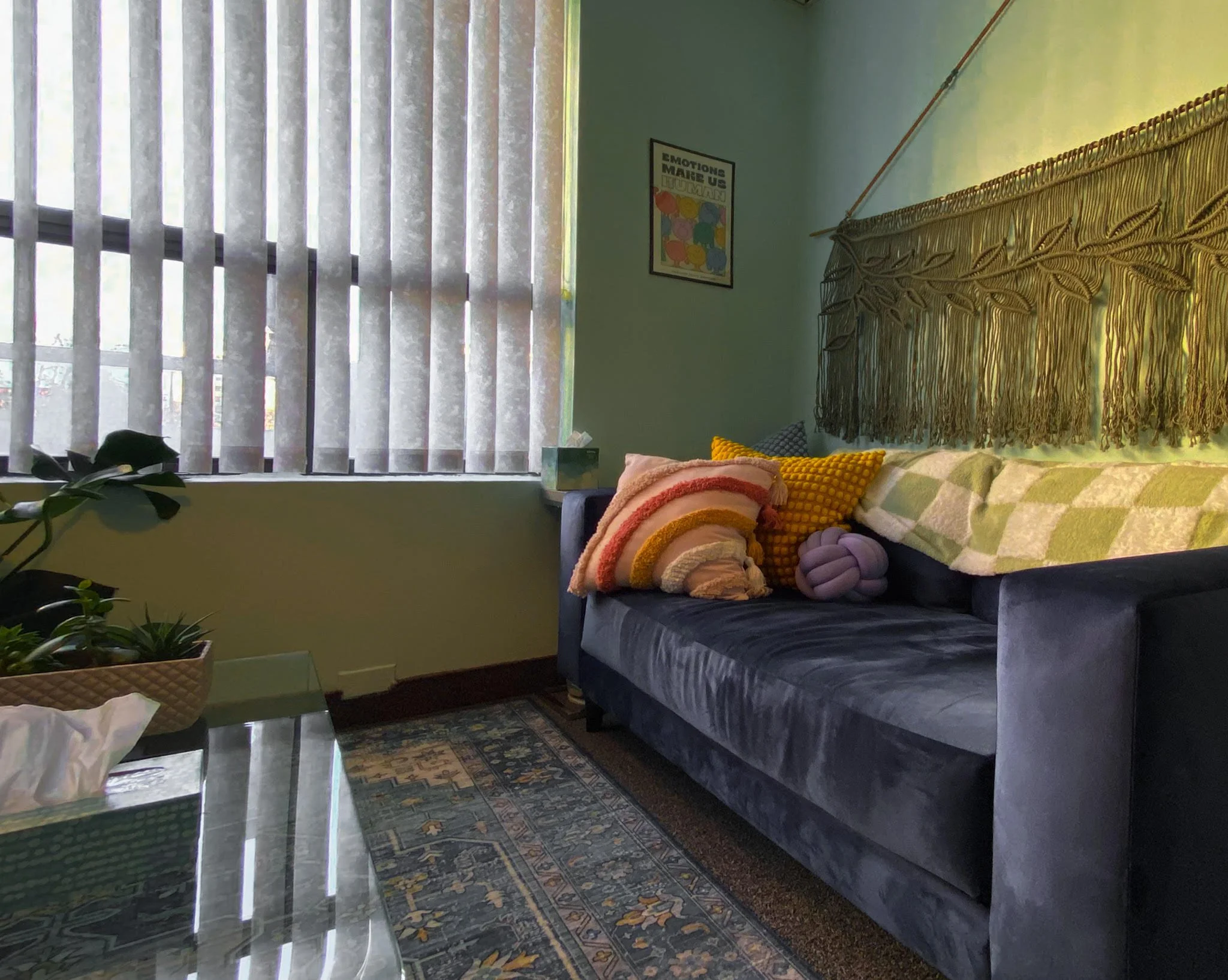 A cozy office with a navy blue velvet sofa decorated with colorful cushions, a macrame wall hanging, a window with vertical blinds, and a patterned rug on the floor.