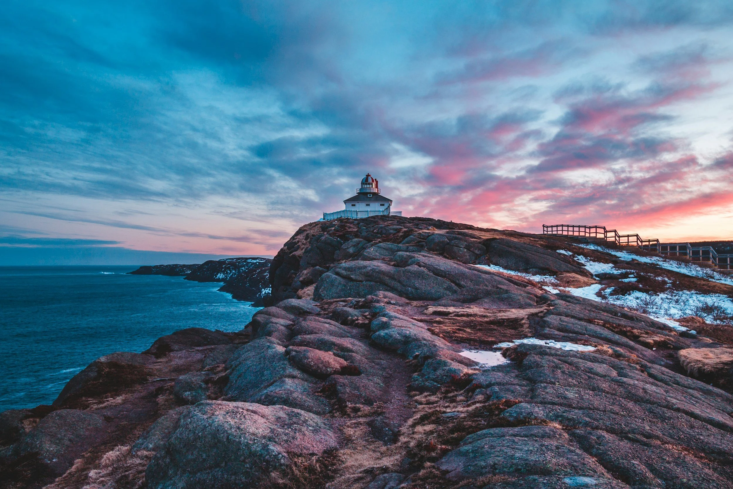 A lighthouse on a rocky coastal cliff during sunset with colorful sky and patches of snow.