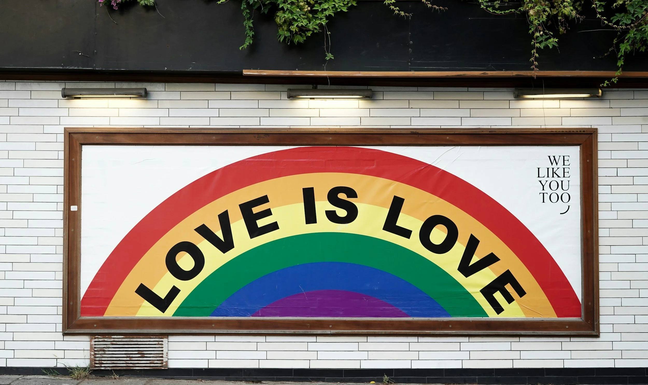 A colorful rainbow poster with the message 'LOVE IS LOVE' and the phrase 'We like you too' in the top right corner, mounted on a white brick wall with three outdoor lights above it.