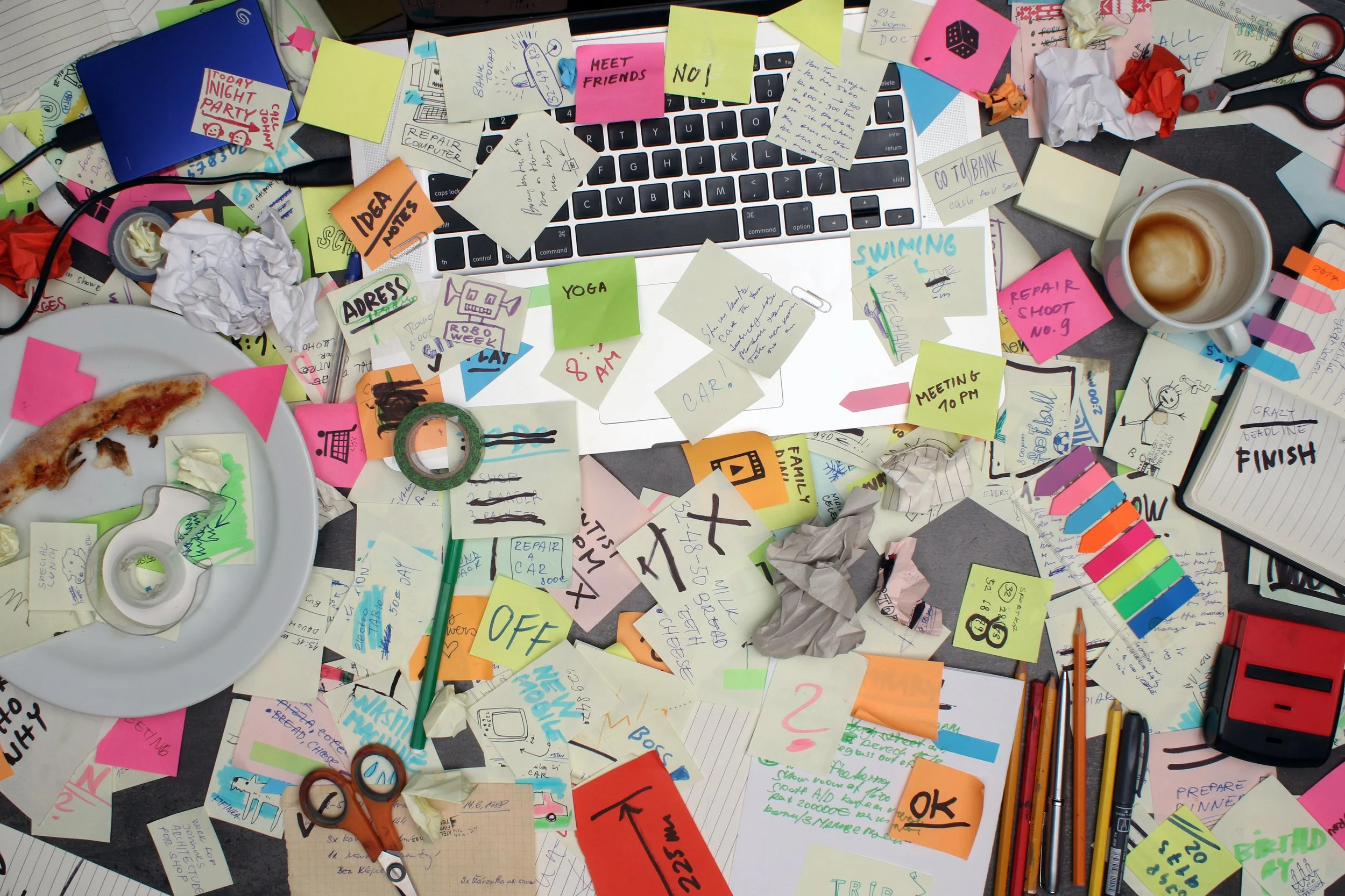 Cluttered, messy desk with scattered sticky notes, crumpled papers, a computer keyboard, a coffee mug, scissors, a slice of pizza on a plate, pens, and various office supplies. What the desk of someone with ADHD might look like.