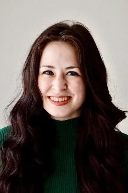 A young woman with long wavy brown hair, smiling, wearing a green turtleneck sweater against a plain white background.