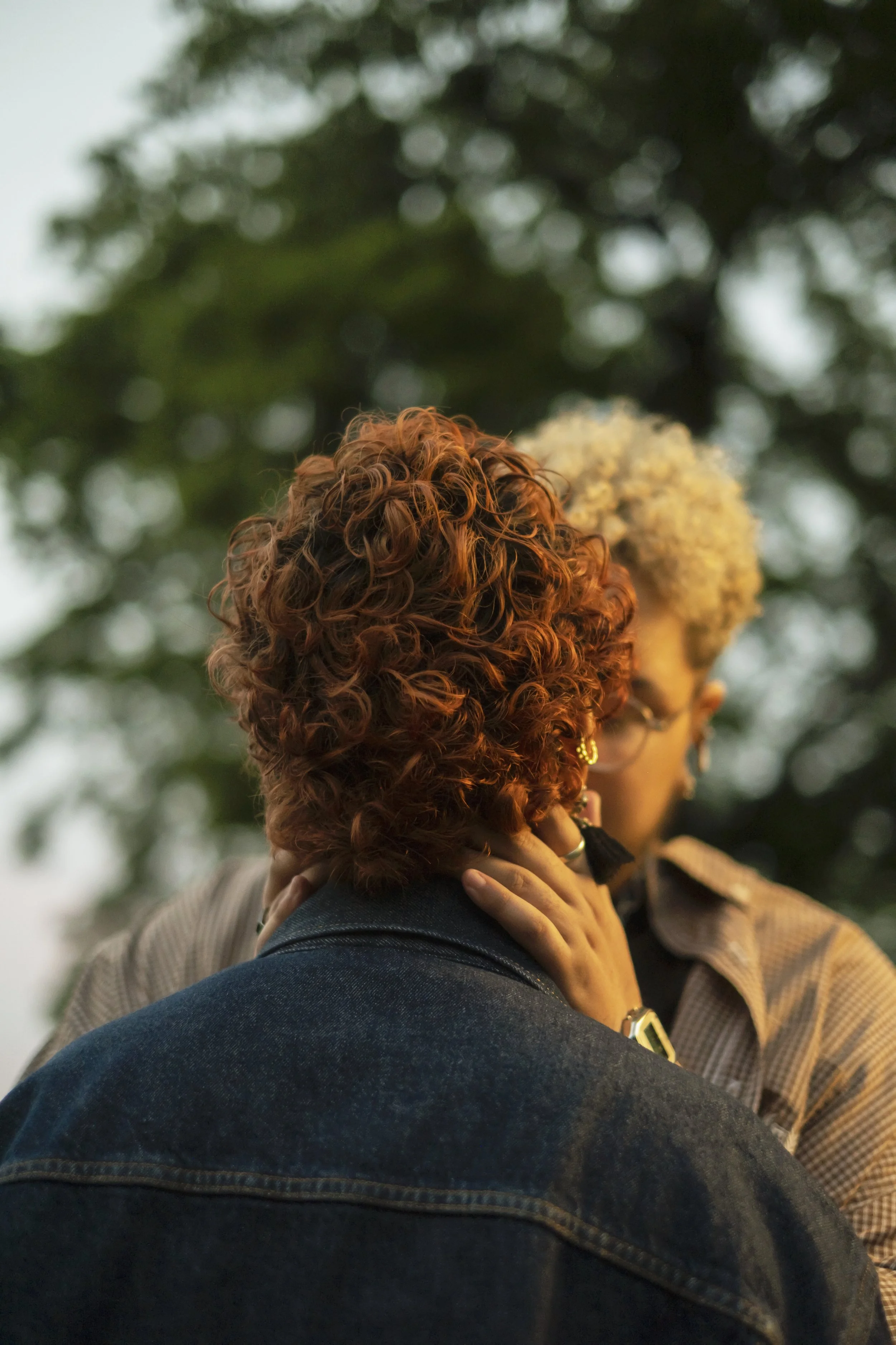 Two people with curly hair, one with red hair and one with blonde hair, are close to each other outdoors, with one person touching the other's neck.