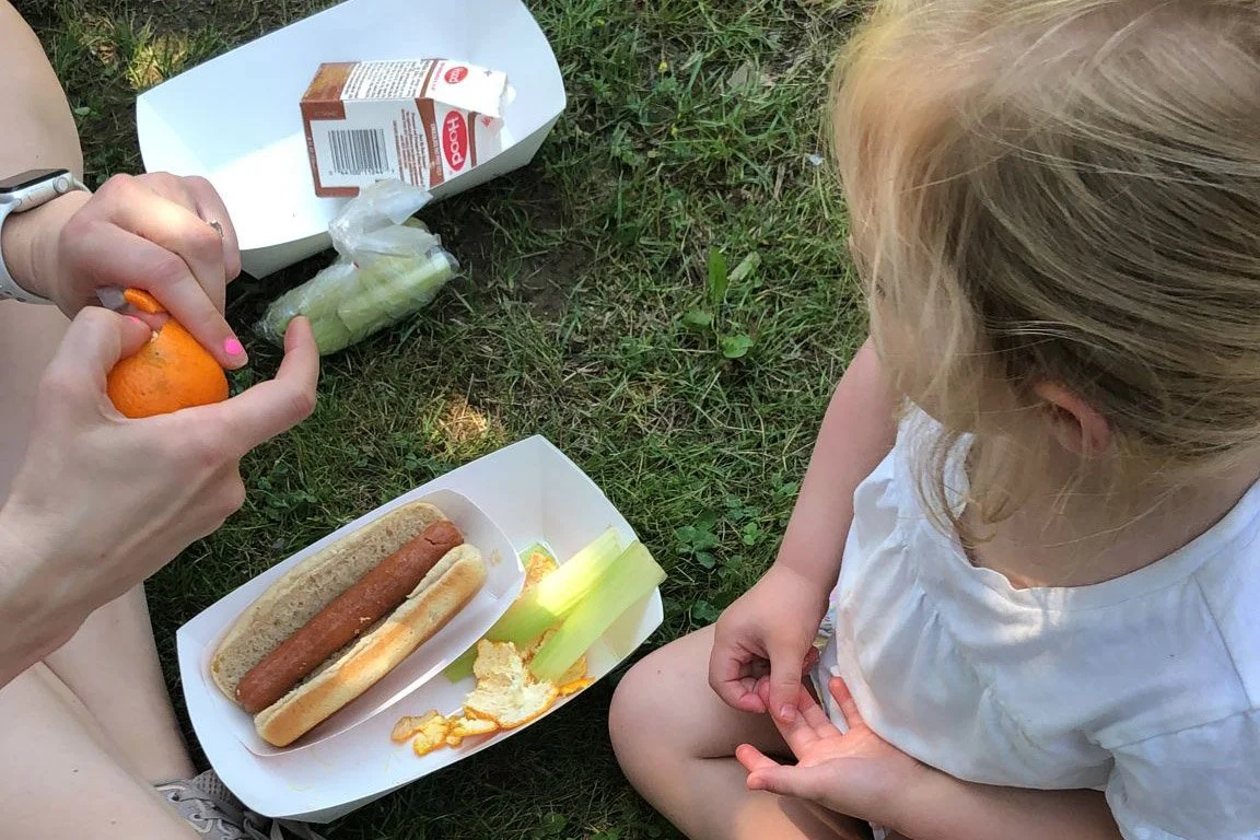 An adult peels a clementine while a child with a white shirt sits nearby on the grass. Two paper trays sit on the grass, with food items like a hot dog and chocolate milk.