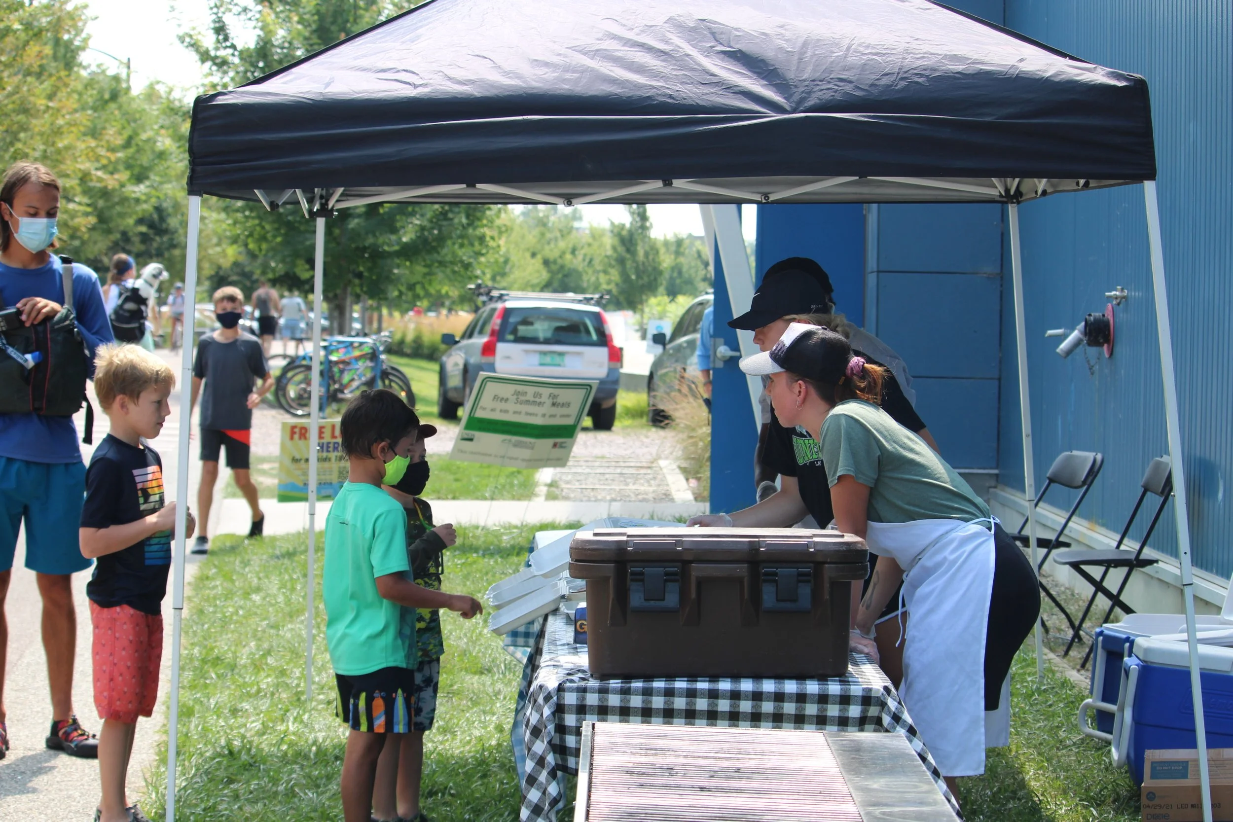 Children in face covering masks stand under a navy blue tent as adults in aprons serve them meals.