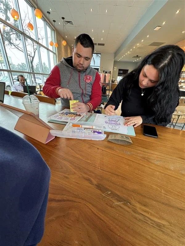 Two young adults sit at a wooden table in a cafe, working on coloring books with colored markers. The cafe has large windows with natural light, hanging round orange lights, and a yellow wall in the background. A drink with a straw is on the table, a