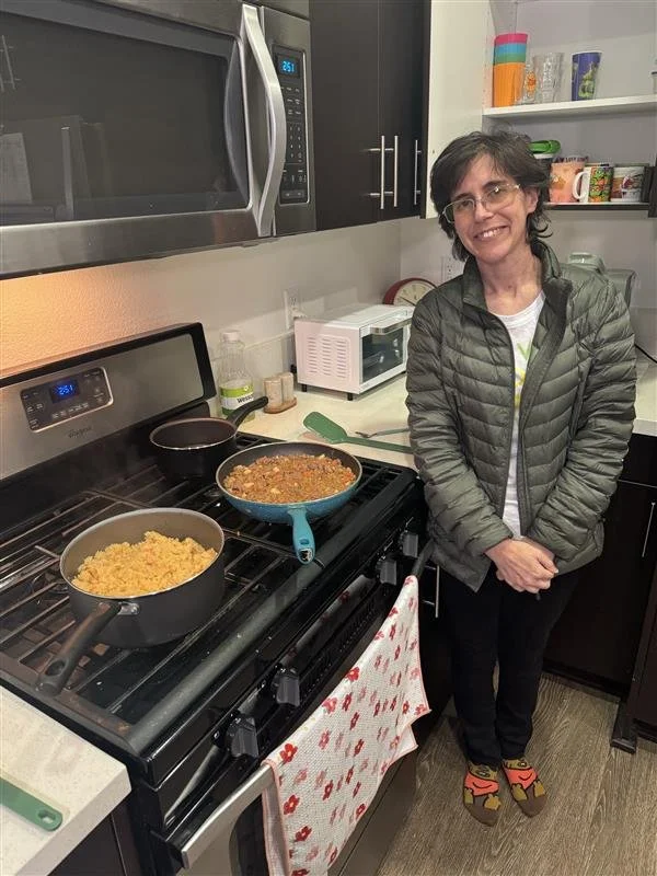 A woman standing in a kitchen next to a stove with two pans, one containing cooked rice and the other a meat and vegetable dish, smiling at the camera.