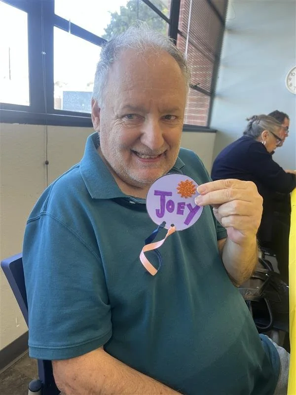 A smiling man holding a purple circular card with the name 'Joey' written on it in purple letters, decorated with an orange flower and a pink ribbon, sitting indoors at a table near large windows.