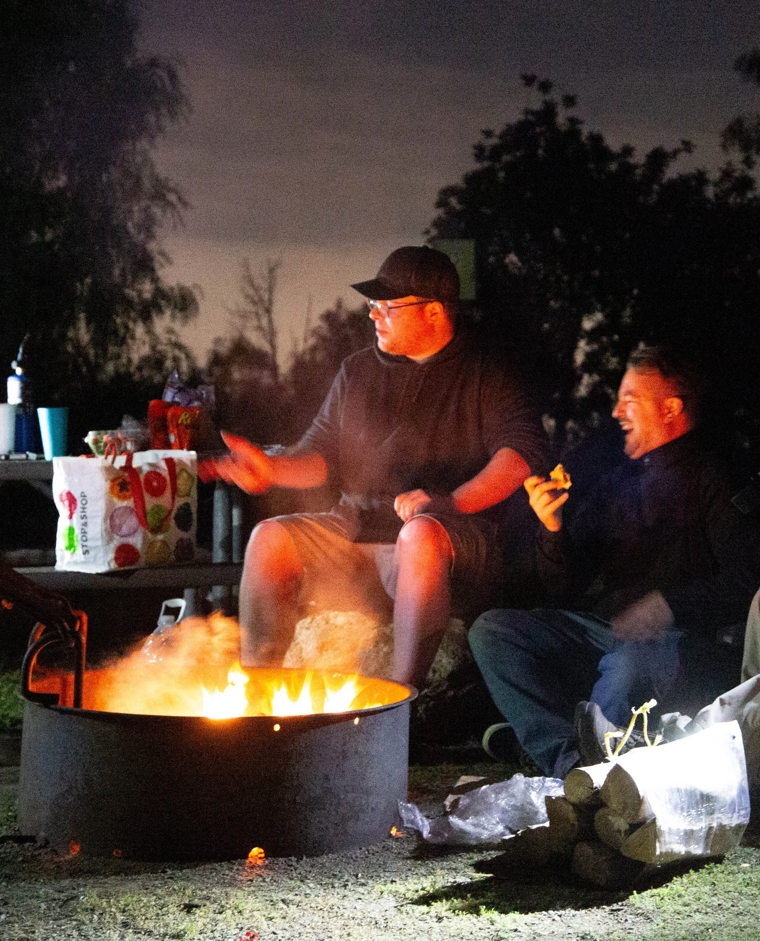Two men sitting around a campfire at night, one of them is holding food and the other is gesturing, with a cooler and snacks nearby.