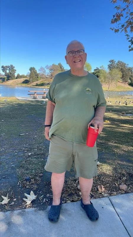 A smiling elderly man with glasses standing outdoors near a pond, holding a red tumbler, wearing a green t-shirt, beige shorts, and black Crocs on a sunny day.