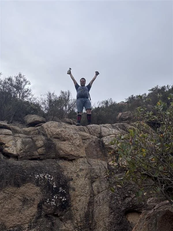 A person standing on top of rocks with arms raised in victory, outdoors with bushes and a cloudy sky in the background.
