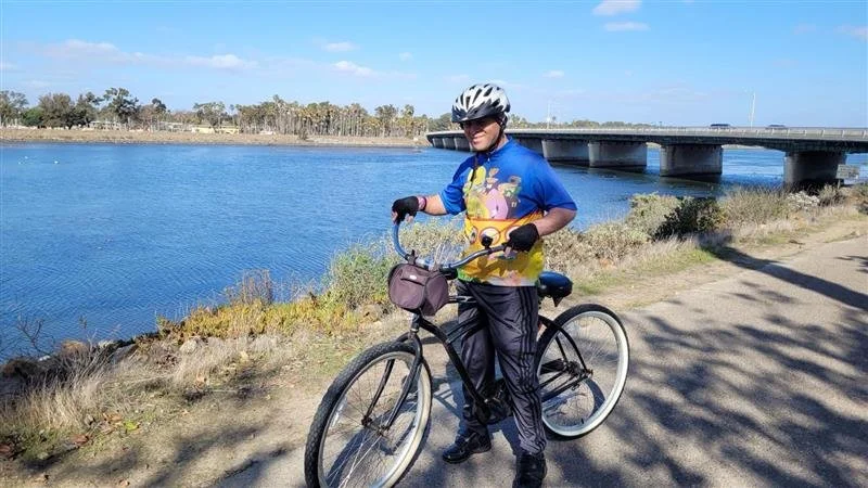 Person wearing a helmet and colorful shirt standing next to a bicycle on a dirt path near a body of water with a bridge in the background.