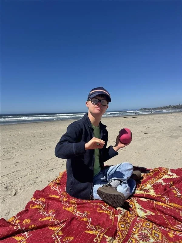 A person sitting on a red patterned blanket at a beach, holding a pink ball, wearing sunglasses, a jacket, and a cap with a blue sky and ocean waves in the background.