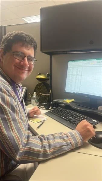 A man with glasses smiling at his desk in an office, working on a computer with a spreadsheet open.