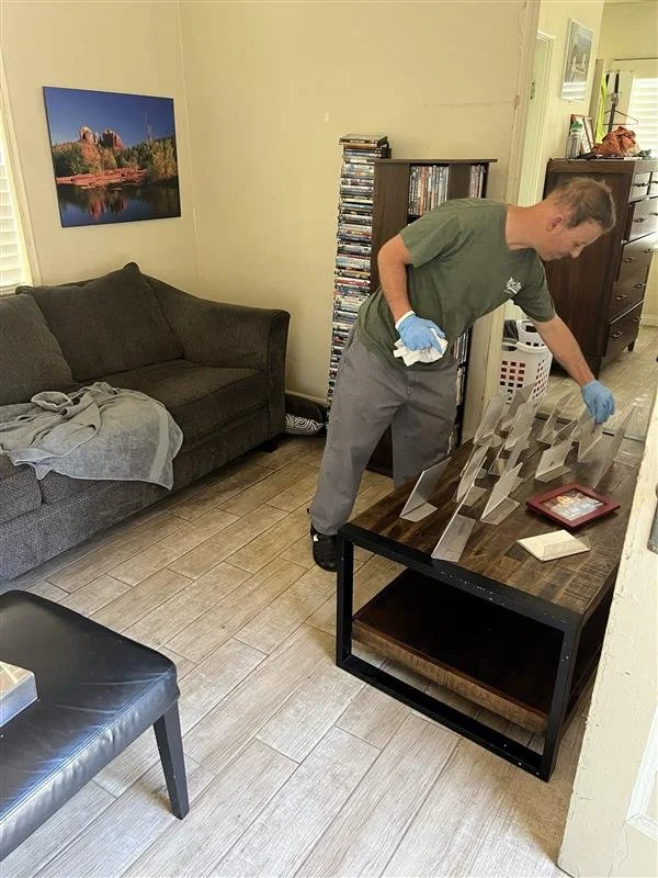 A man with gloves and a cloth cleaning or inspecting a series of glass or acrylic display stands on a wooden table in a living room.