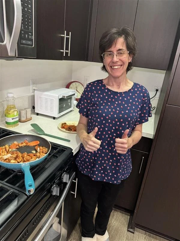 A woman with glasses and short dark hair standing in a kitchen, smiling and giving two thumbs up. There is a frying pan with cooked food on the stove, a microwave, a plate with food, and some bottles and containers on the countertop.