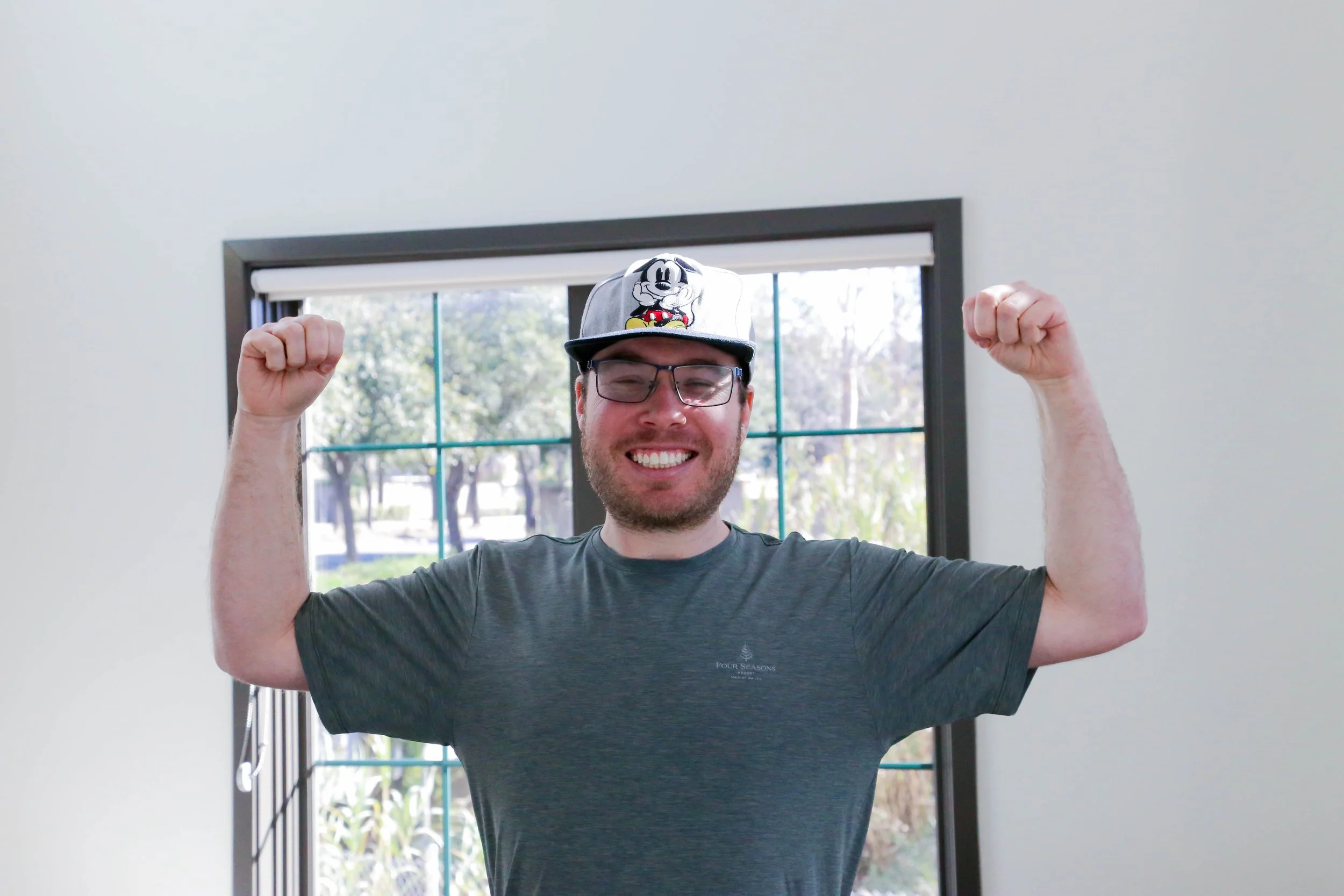 Man with glasses smiling and flexing muscles, wearing a Mickey Mouse cap in front of a window with trees outside.